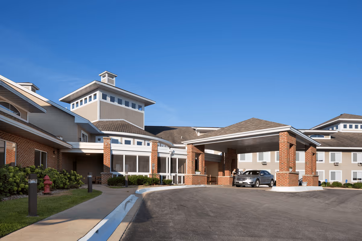 Exterior view of Thomas Grove Assisted Living and Memory Care facility showing a large covered entrance with brick pillars, a driveway, and a parked car. The building has multiple windows and a combination of brick and siding under a clear blue sky.