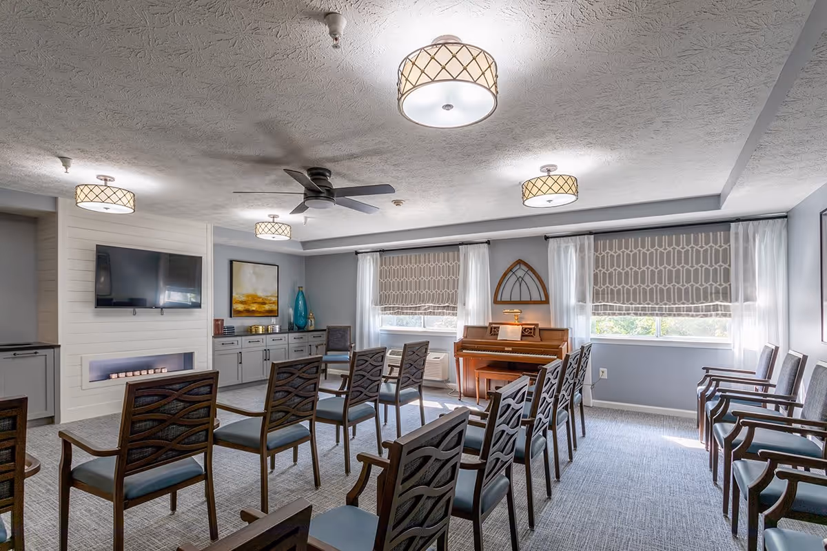 A well-lit common room with multiple rows of wooden chairs facing a wall-mounted flat screen TV and a modern electric fireplace. The room features a piano against the far wall, two windows with patterned Roman shades and sheer curtains, a ceiling fan, and several ceiling lights with decorative shades. The walls are painted light gray and there is a sideboard with decorative items and artwork.