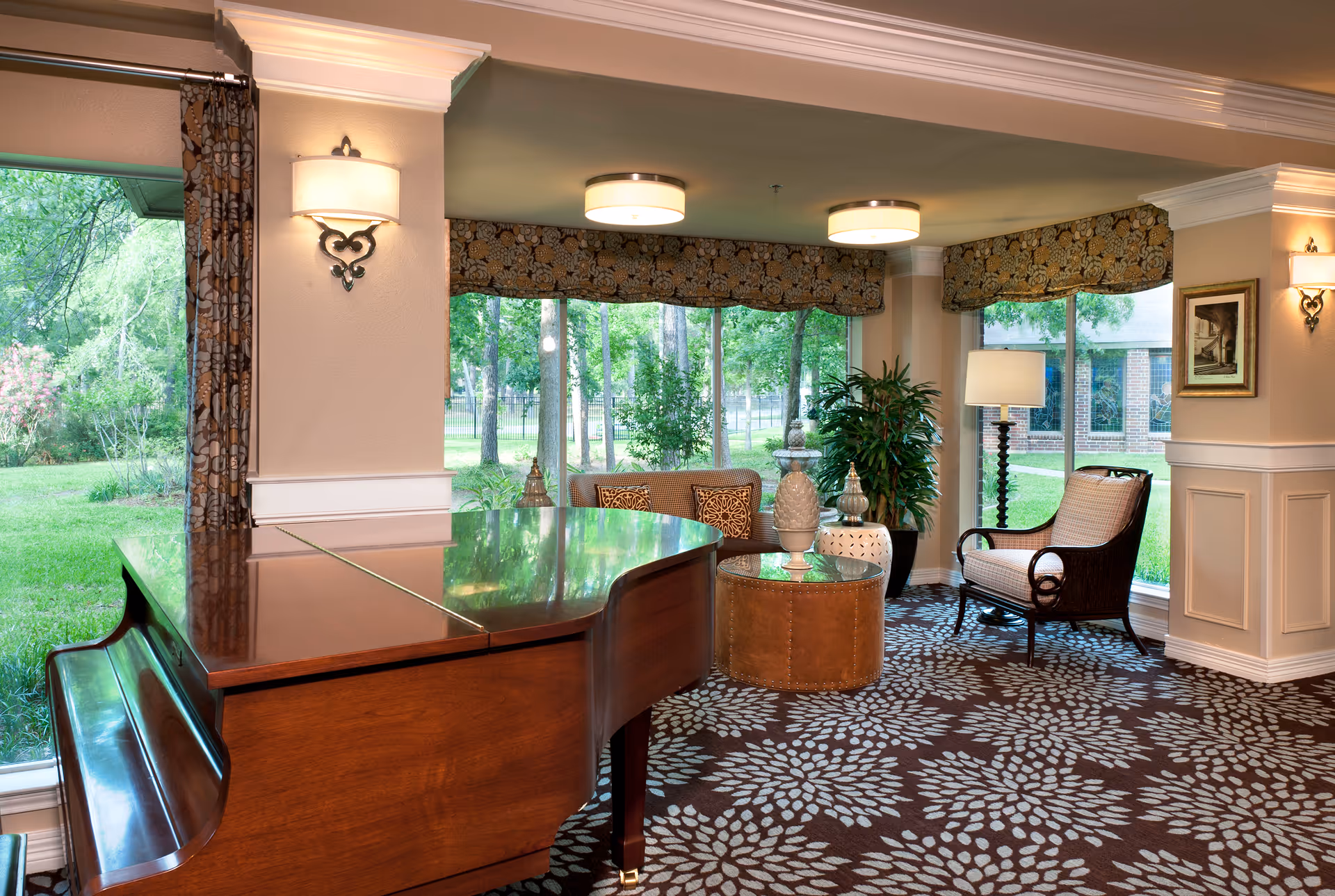 A cozy sitting area in a retirement community featuring a polished wooden grand piano, a round leather ottoman, a cushioned armchair, a small sofa with decorative pillows, a floor lamp, and large windows with floral valances overlooking a green outdoor space with trees and grass.