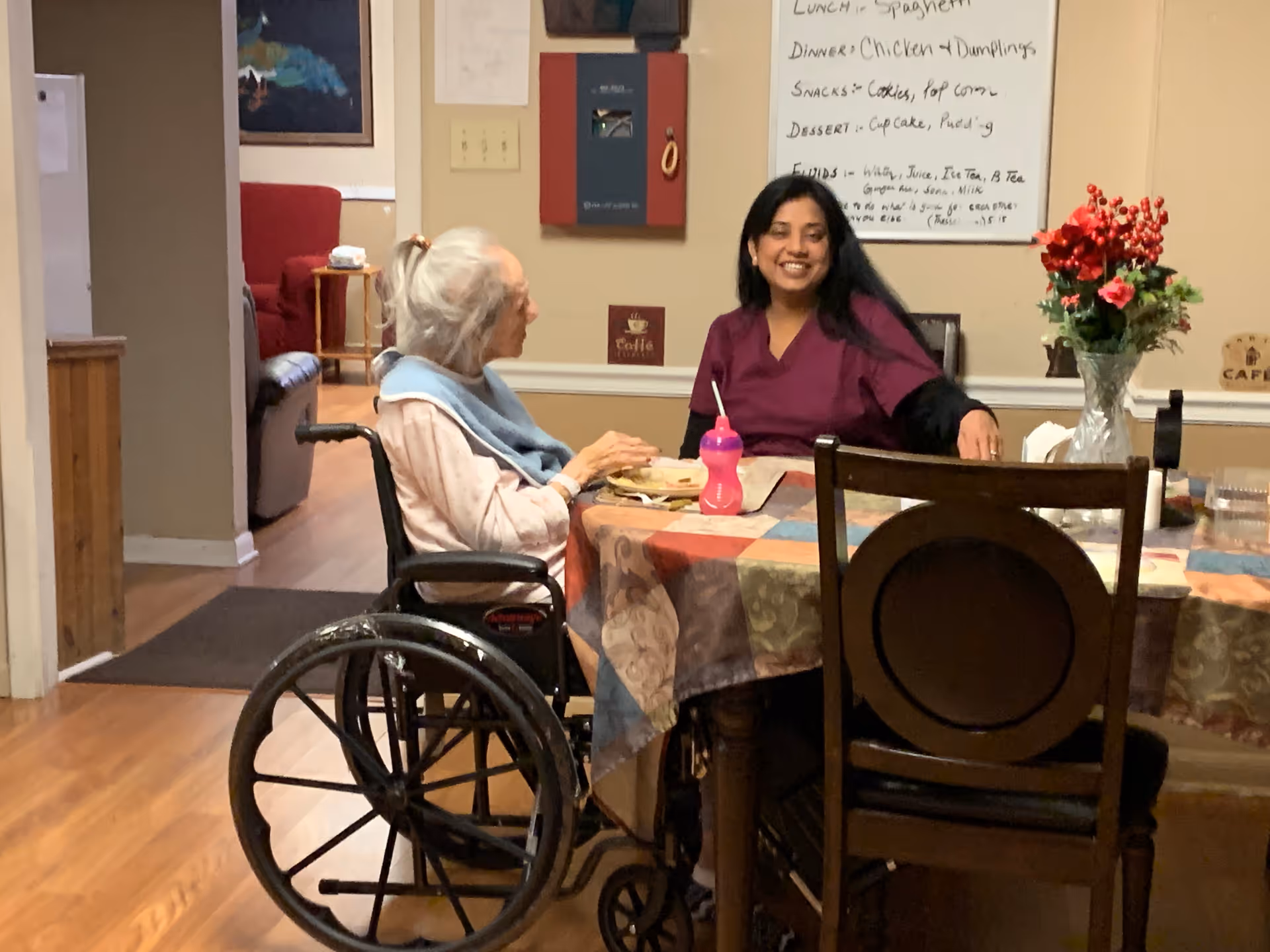 An elderly woman in a wheelchair and a caregiver in a maroon uniform sitting at a dining table covered with a colorful tablecloth, smiling and engaging in conversation. The room has wooden flooring, a vase with red flowers on the table, and a whiteboard with meal information on the wall behind them.