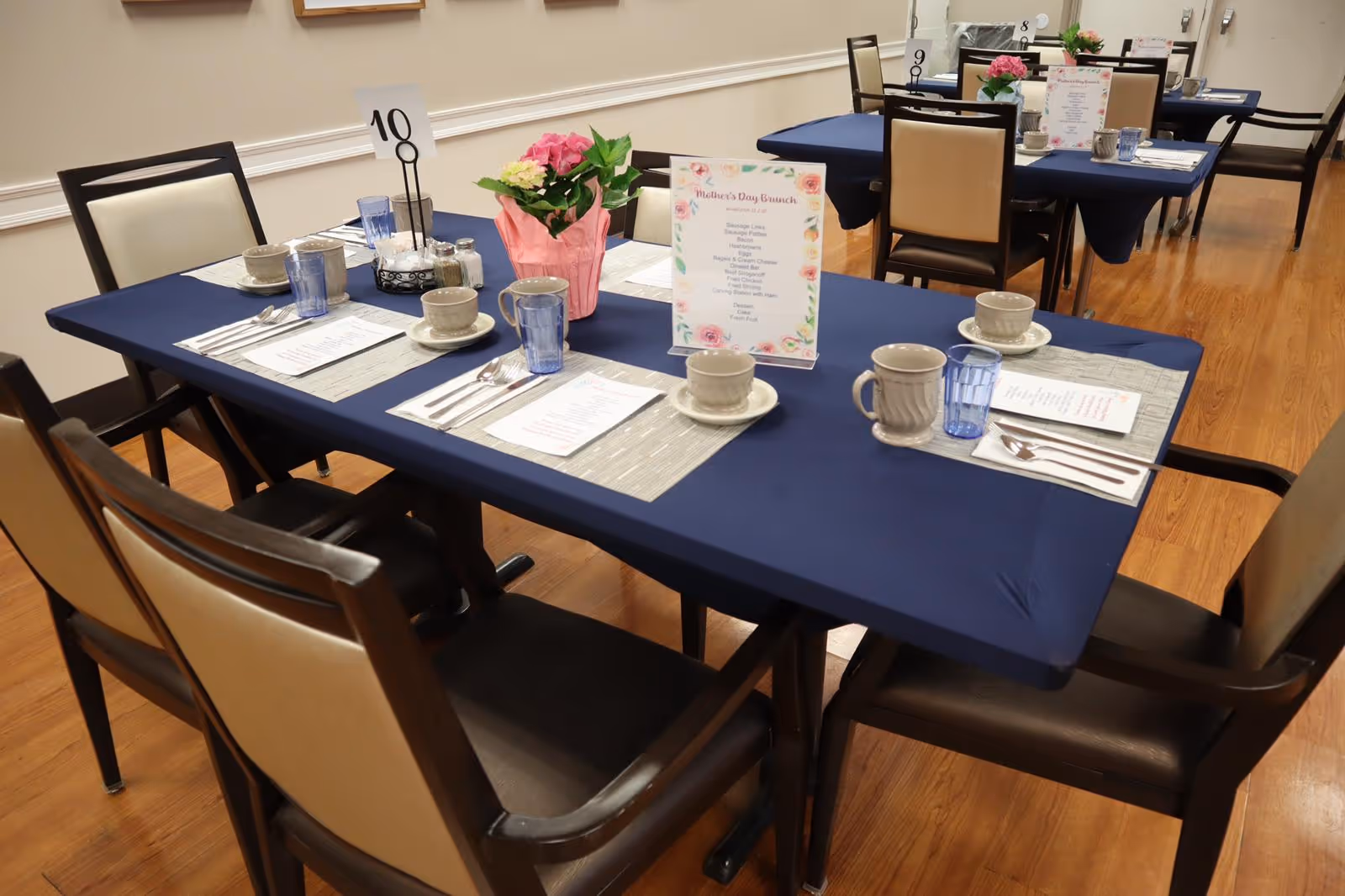 A dining room with tables covered in navy blue tablecloths, set with placemats, menus, cups, glasses, and utensils. Each table has a small flower arrangement and a table number. The floor is wooden, and the room has a simple, clean design.