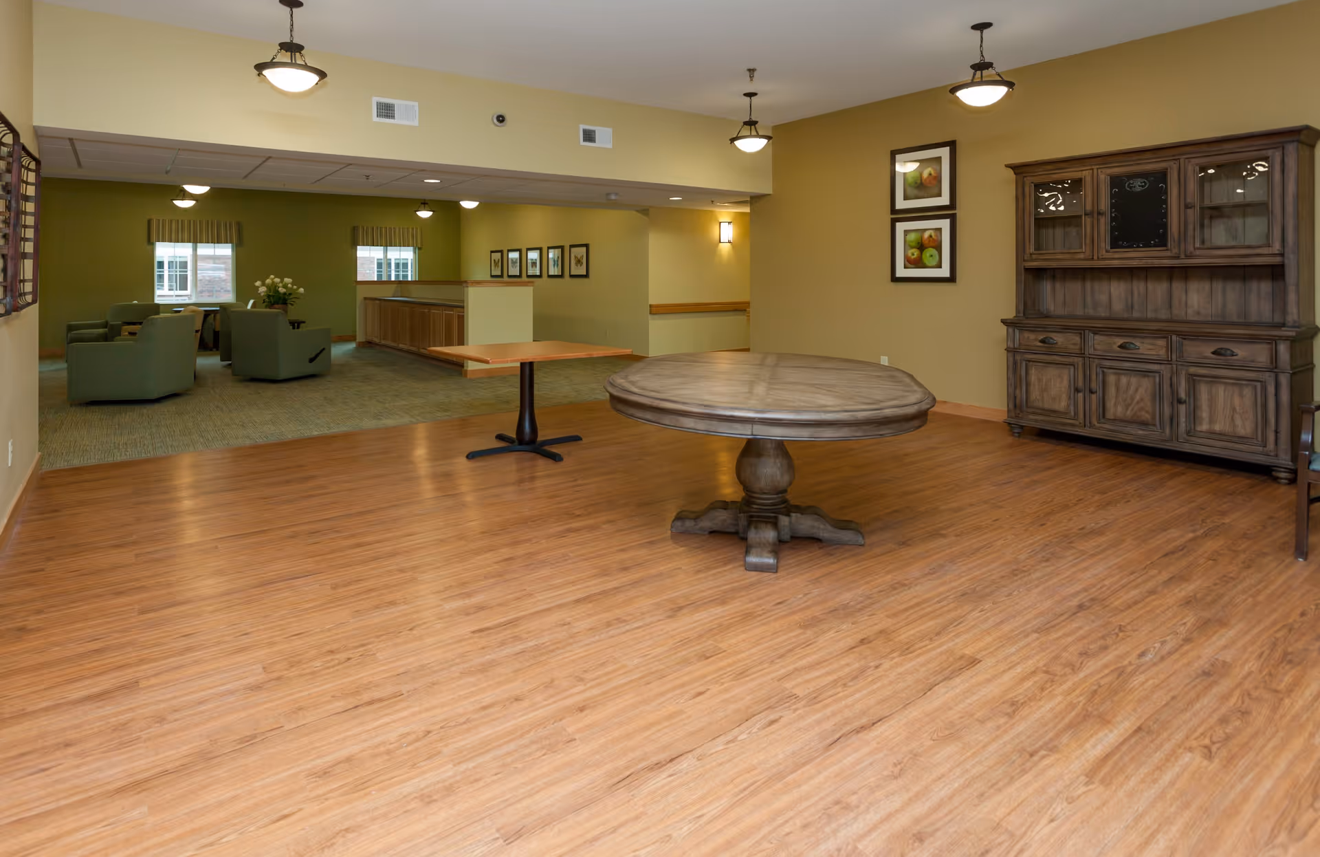 A spacious interior room with wooden flooring and beige walls. The room features a large round wooden table in the center, a smaller rectangular table, and a wooden cabinet with glass doors on the right. In the background, there is a carpeted seating area with green armchairs, a window with striped curtains, and framed pictures on the walls. Ceiling lights illuminate the space.