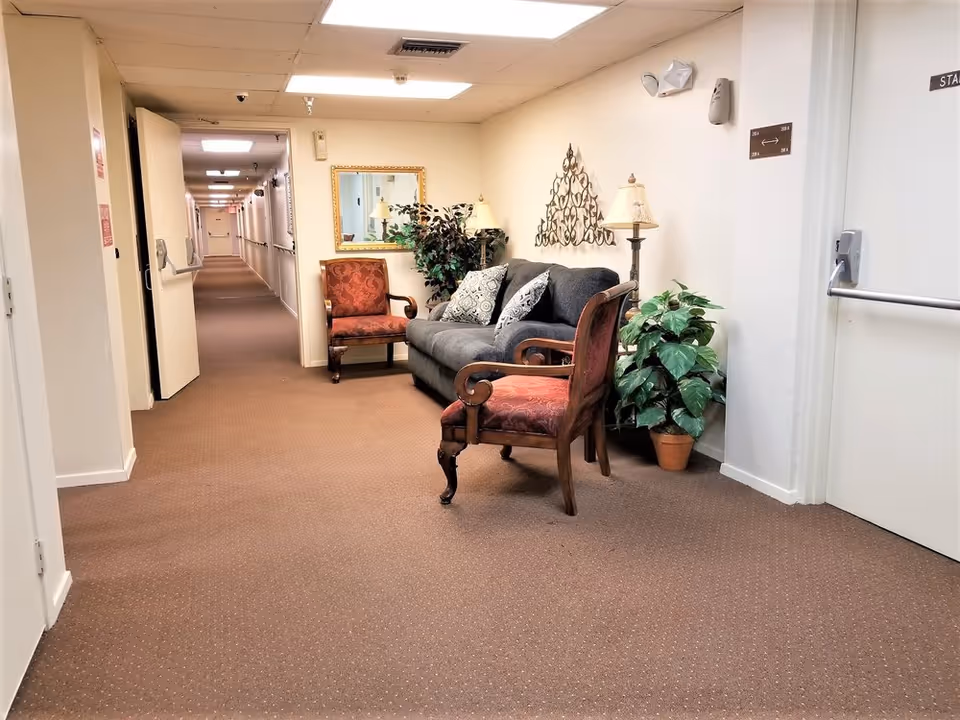 A hallway in a senior living facility with brown carpet and white walls. There is a seating area with a gray couch, two red upholstered chairs, decorative pillows, a floor lamp, a potted plant, a wall mirror, and wall art. The hallway extends into the distance with several doors and handrails along the walls.
