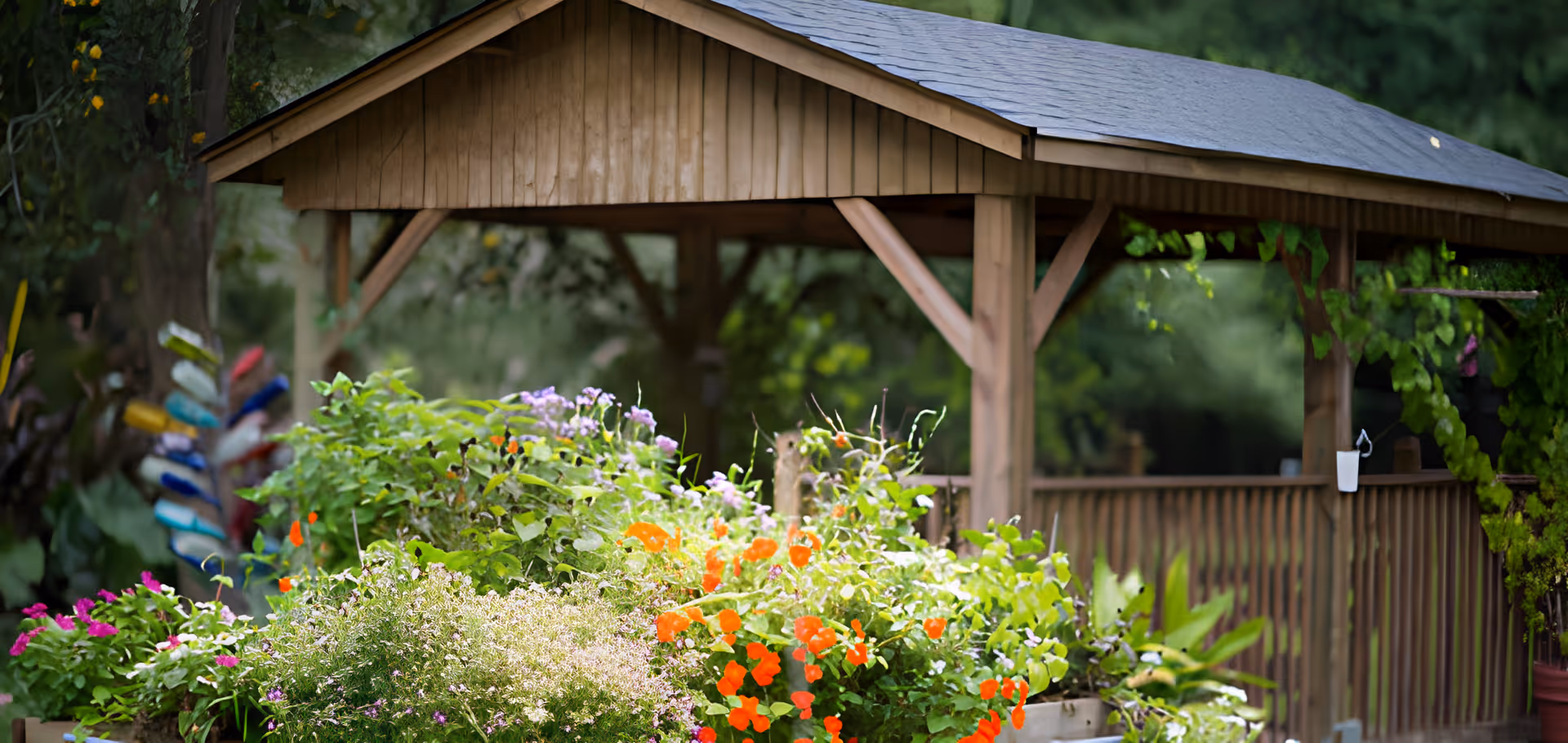A wooden gazebo surrounded by a vibrant garden with various colorful flowers including orange, purple, and pink blooms, set in a lush green outdoor area.