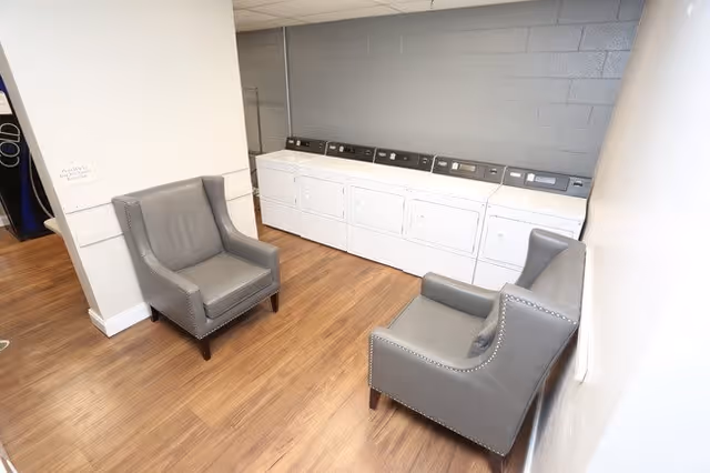 A laundry room with six white washing machines and dryers lined up against a gray wall. Two gray upholstered armchairs with nailhead trim are placed on a wooden floor, facing each other near the machines. The room has white walls and a ceiling with fluorescent lighting.