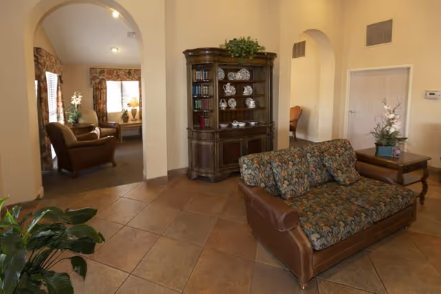 A cozy living area in a senior care facility featuring a floral-patterned sofa with leather arms, a wooden display cabinet with decorative plates and books, and an adjacent room with armchairs and a window with floral curtains. The space has tiled flooring, arched doorways, and potted plants adding greenery.