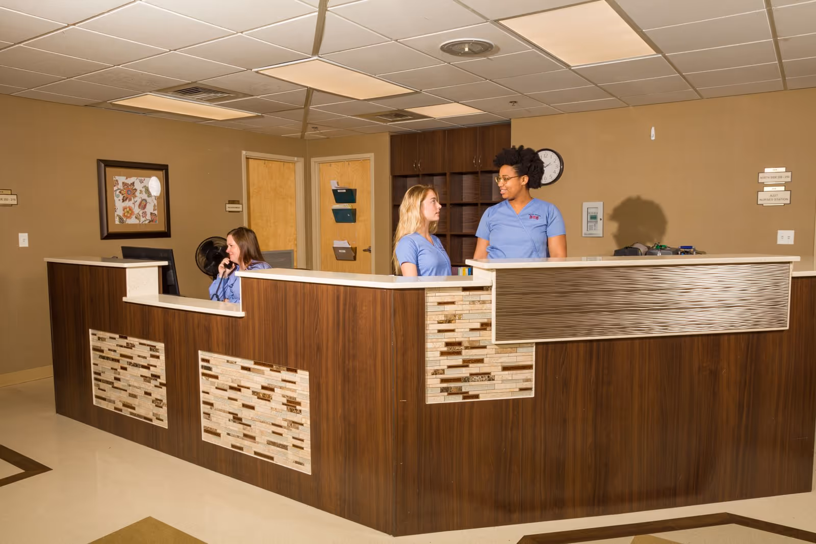 A reception area inside a healthcare facility with a wooden front desk featuring decorative tile accents. Three staff members in blue uniforms are present; one is seated and talking on the phone, while two others are standing and conversing. The background includes two closed wooden doors, a wall clock, and some wall-mounted organizers.