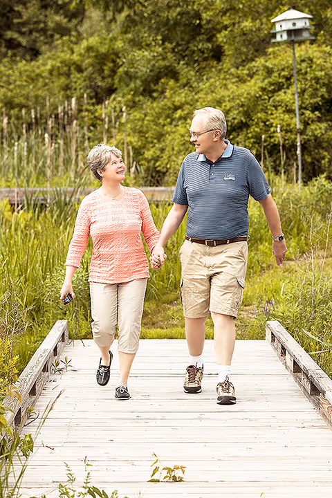 An older couple holding hands and walking on a wooden boardwalk surrounded by lush green vegetation and trees. The woman is wearing a coral long-sleeve top and light-colored pants, while the man is wearing a blue striped polo shirt and beige shorts. Both are smiling and looking at each other.