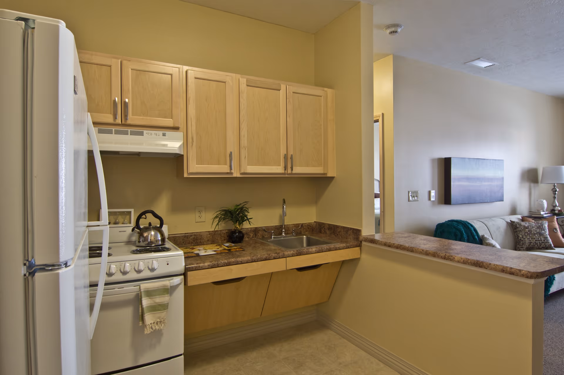 A small kitchen area with light wood cabinets, a white refrigerator, a white stove with a kettle on it, and a countertop with a sink. The kitchen opens into a living room with a beige couch, decorative pillows, a lamp, and a wall-mounted artwork.