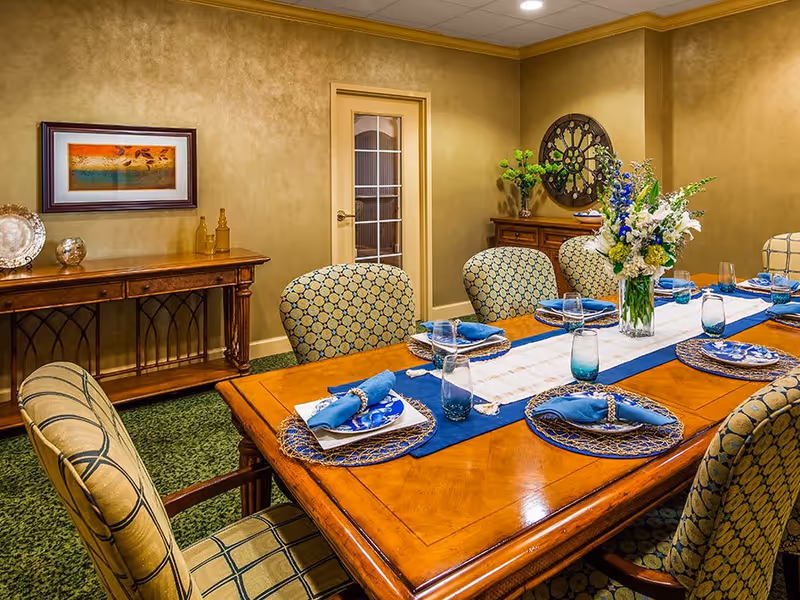 A formal dining room with a wooden table set for six people. The table has blue and white plates, blue napkins, clear drinking glasses, and a floral centerpiece with white and blue flowers. The chairs around the table have patterned upholstery. The room has beige walls, a green carpet, a wooden sideboard with decorative items, and a framed artwork on the wall.