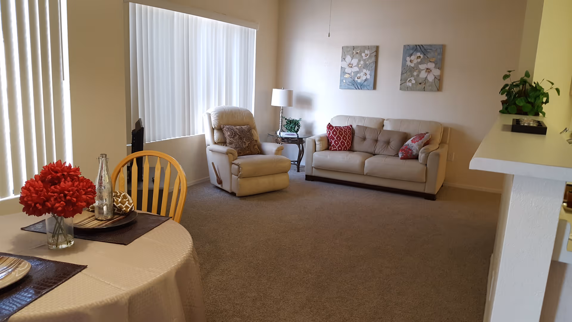 Bright living room and dining area with a beige sofa, recliner, round table with red flowers, and vertical blinds.