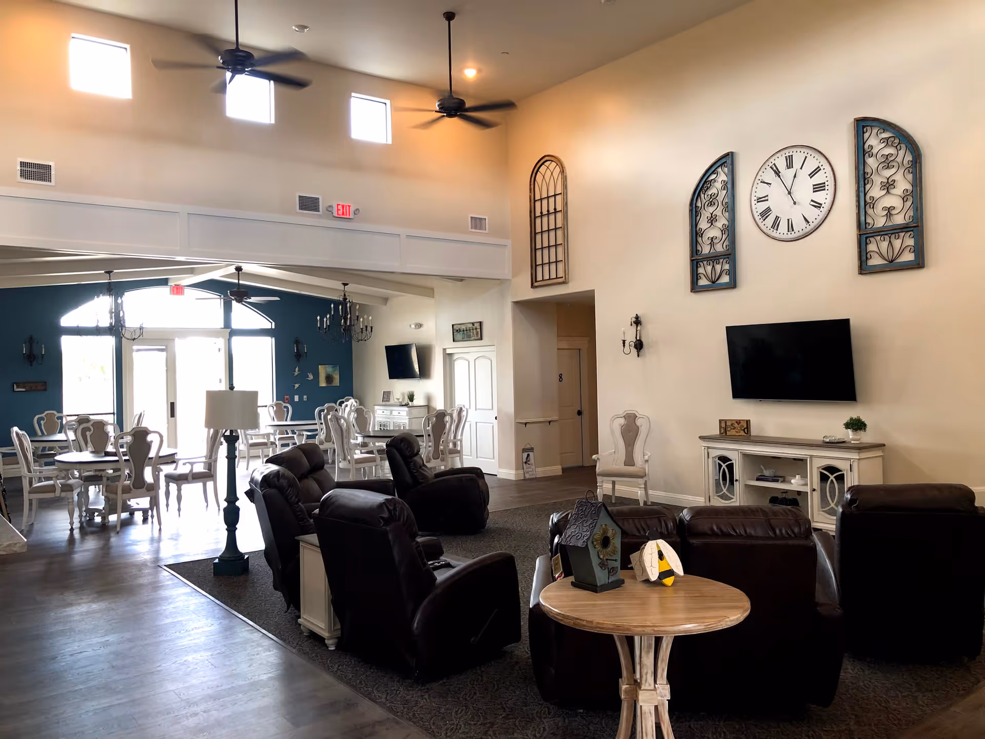 A spacious and well-lit common area in an assisted living facility featuring a seating area with dark leather recliners arranged around a small wooden table with decorative items. A wall-mounted TV is above a white cabinet, and a large clock with Roman numerals is on the wall. In the background, there is a dining area with white chairs and tables, ceiling fans, and large windows letting in natural light.