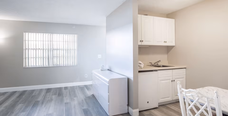 Bright studio-style interior with a small kitchenette, dining table, dresser, and a window with vertical blinds.