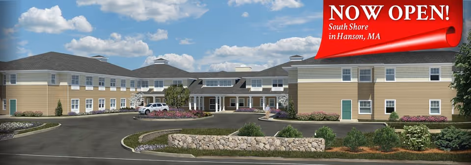 Front exterior of a two-story assisted living building with a circular driveway and landscaping, topped by a red "NOW OPEN" banner.