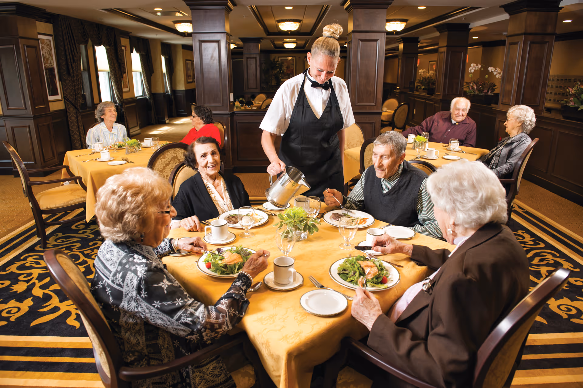 Elderly residents sit around a dining table eating salads while a server pours coffee in an elegant dining room.