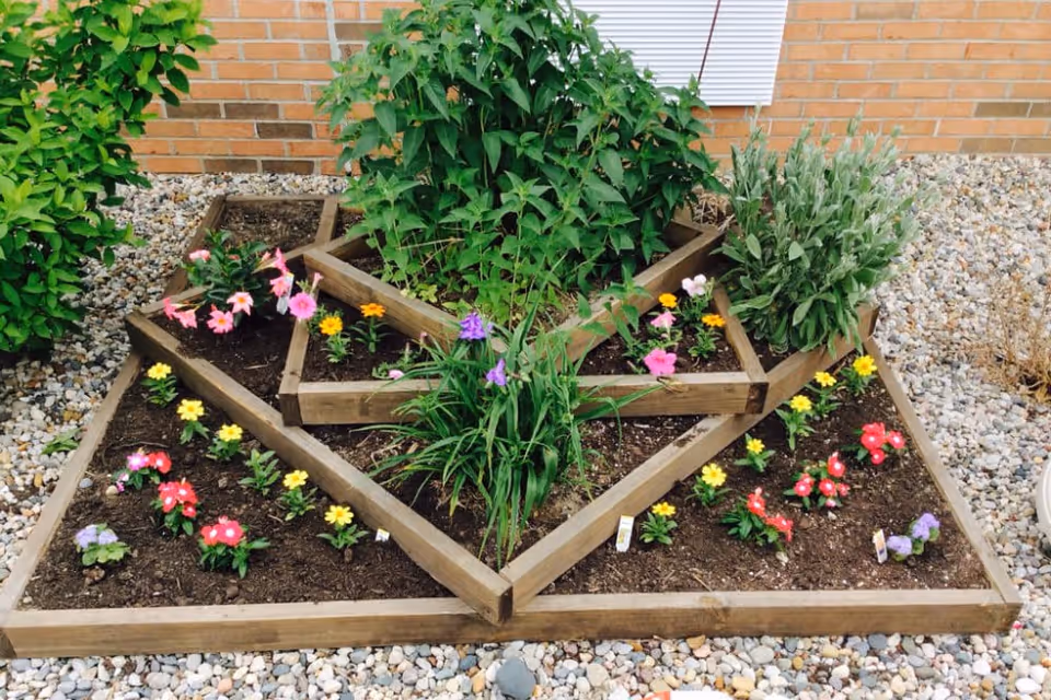 A geometric raised garden bed with various colorful flowers and green plants, surrounded by small pebbles and set against a brick wall.