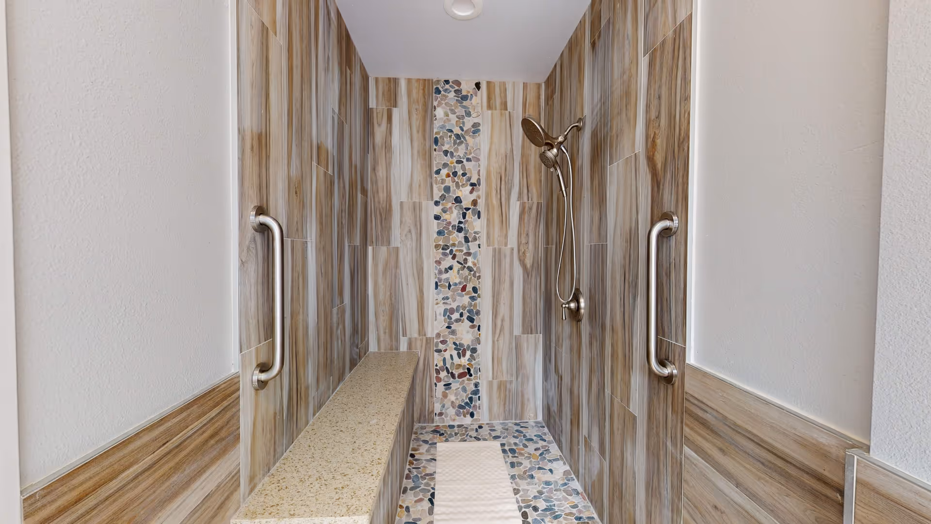 Walk-in tiled shower with a built-in bench, stainless steel grab bars, a handheld showerhead, and a pebble-tile accent strip.