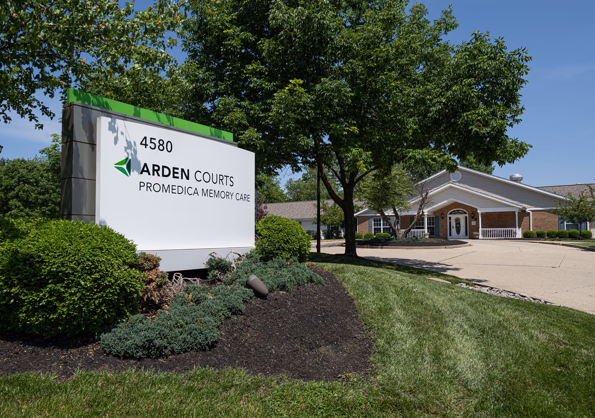 Exterior view of Arden Courts - ProMedica Memory Care Community building with a large sign displaying the facility name and address 4580, surrounded by green bushes and trees under a clear blue sky.