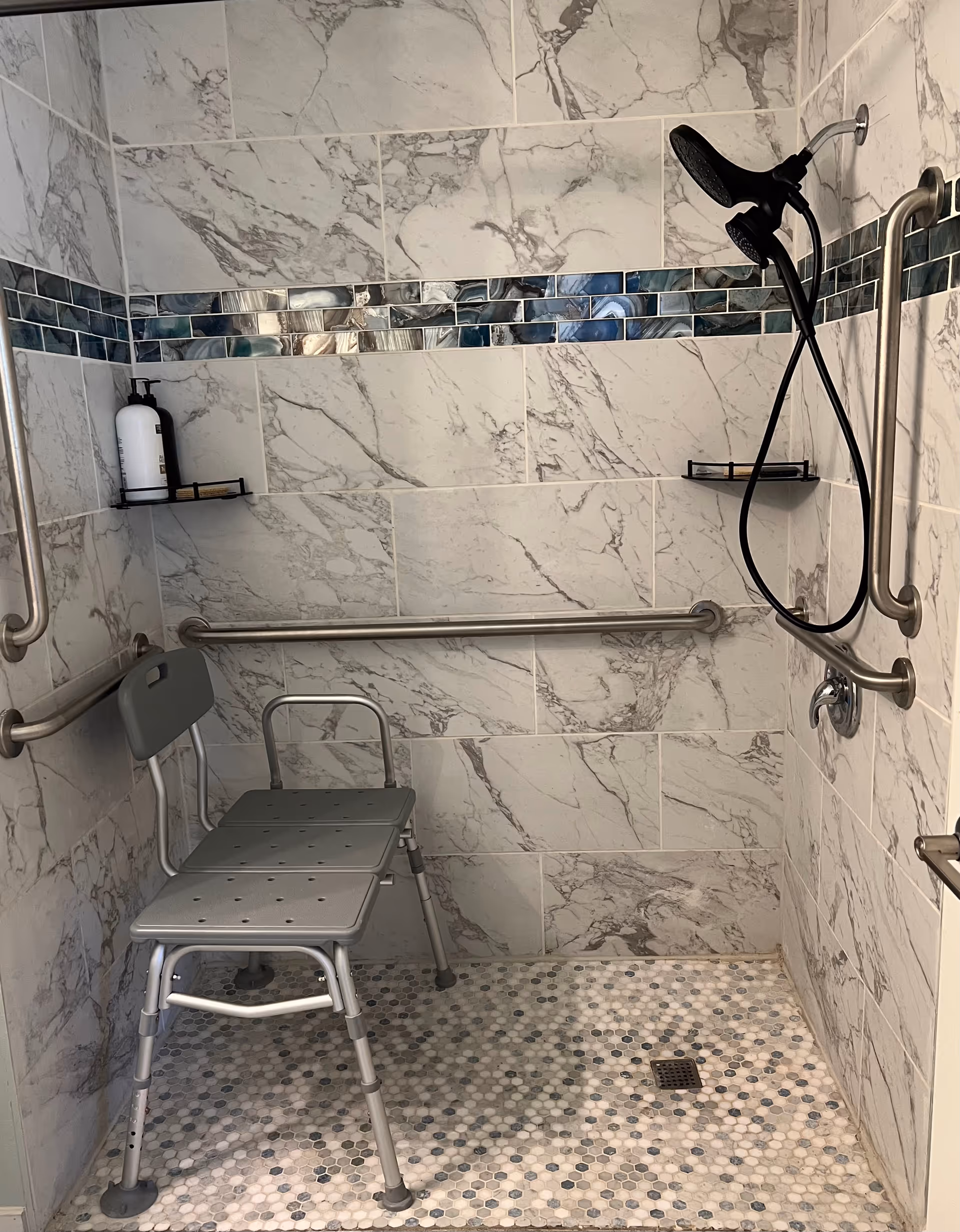 Accessible shower area with marble-patterned wall tiles and a strip of blue and gray mosaic tiles. The shower has a handheld showerhead attached to the wall, multiple stainless steel grab bars, a gray shower chair with backrest and armrest, and a small corner shelf holding a white bottle and a soap bar.