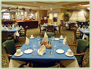 A dining room with multiple tables set for a meal. Each table is covered with a blue tablecloth and white napkins folded in a decorative manner. The tables have plates, cups, glasses, and silverware arranged neatly. The room has carpeted floors, green cushioned chairs, and a buffet area with floral arrangements in the background.