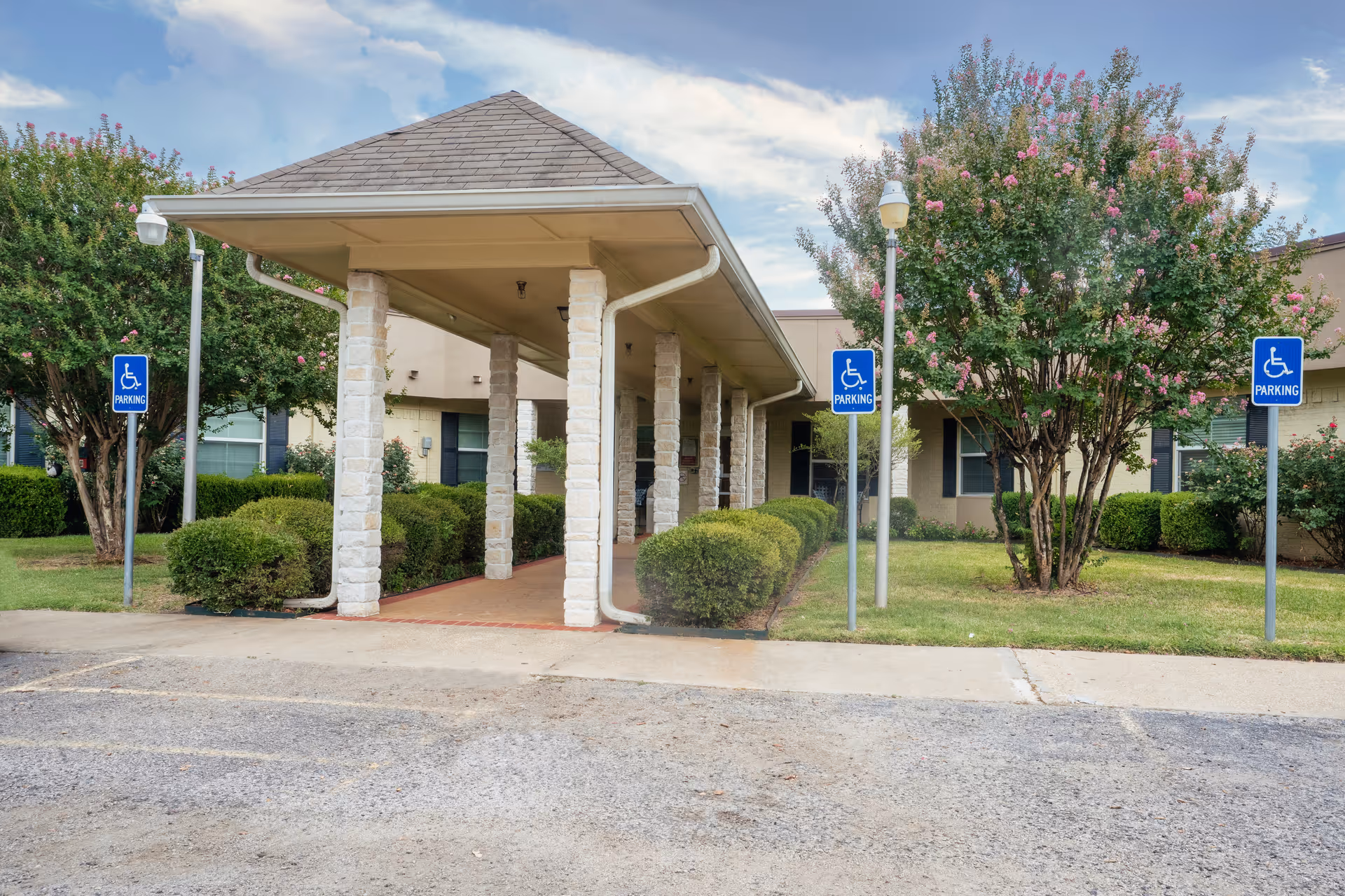 Entrance of Country Meadows Nursing & Rehabilitation Center showing a covered walkway supported by stone pillars, surrounded by green bushes and trees with pink flowers. There are three blue handicap parking signs visible near the entrance.
