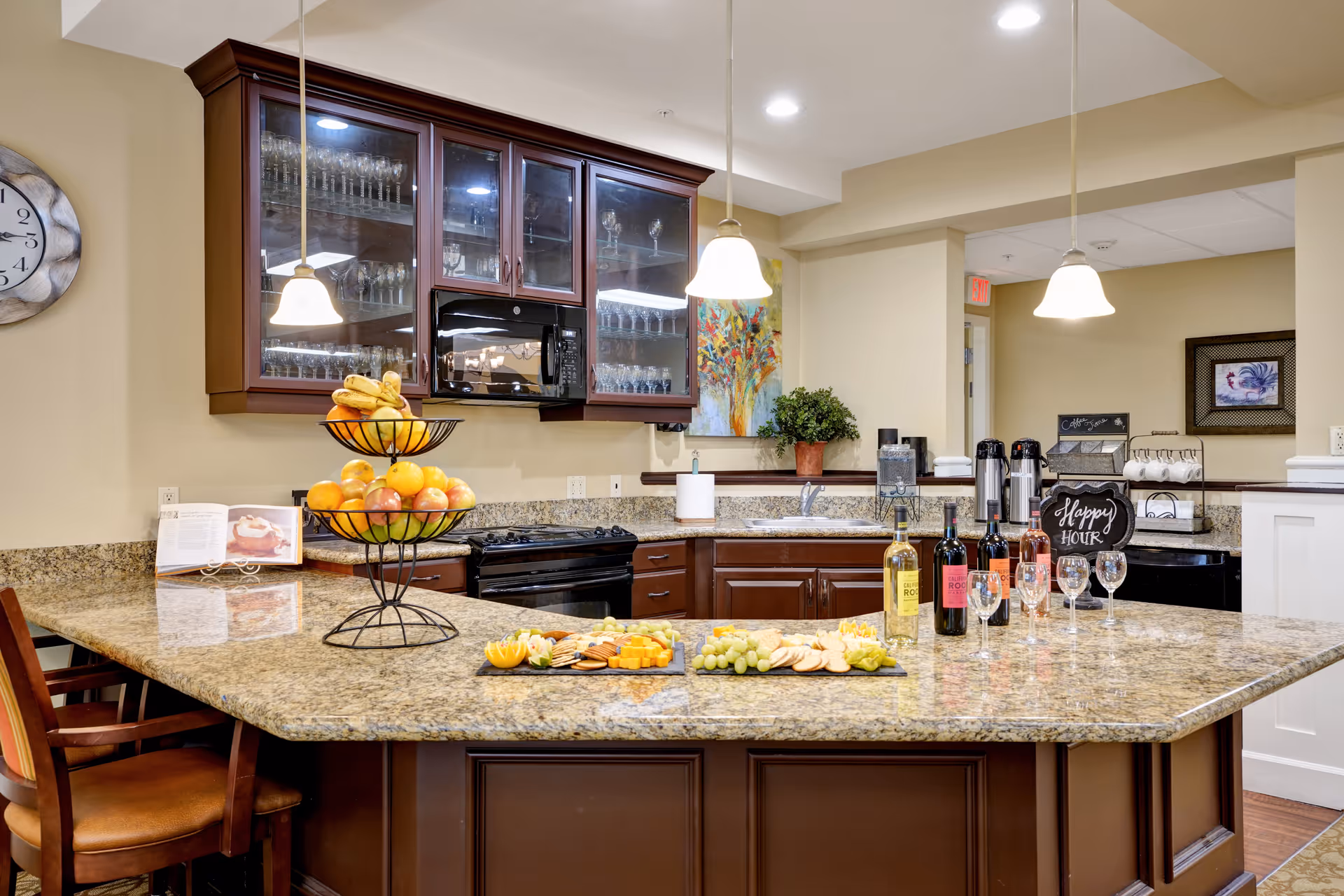 A well-lit kitchen area with granite countertops and dark wooden cabinets. The countertop features a tiered fruit basket with bananas and oranges, a platter with cheese, crackers, and grapes, and several bottles of wine with wine glasses. There are pendant lights hanging above the counter, a microwave mounted above the stove, and glassware displayed in the upper cabinets. A small chalkboard sign reads 'Happy Hour'.
