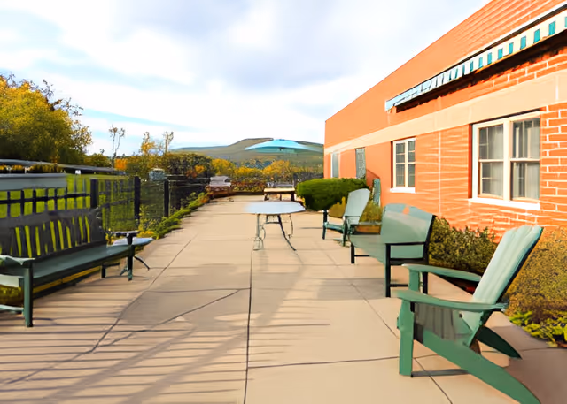 Outdoor patio area with green benches and chairs along a paved walkway next to a brick building. There are bushes and a black metal fence on the left side, with trees and hills visible in the background under a partly cloudy sky.