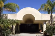 Entrance of a building with an arched architectural feature above the doorway, flanked by palm trees and other greenery under a clear blue sky.
