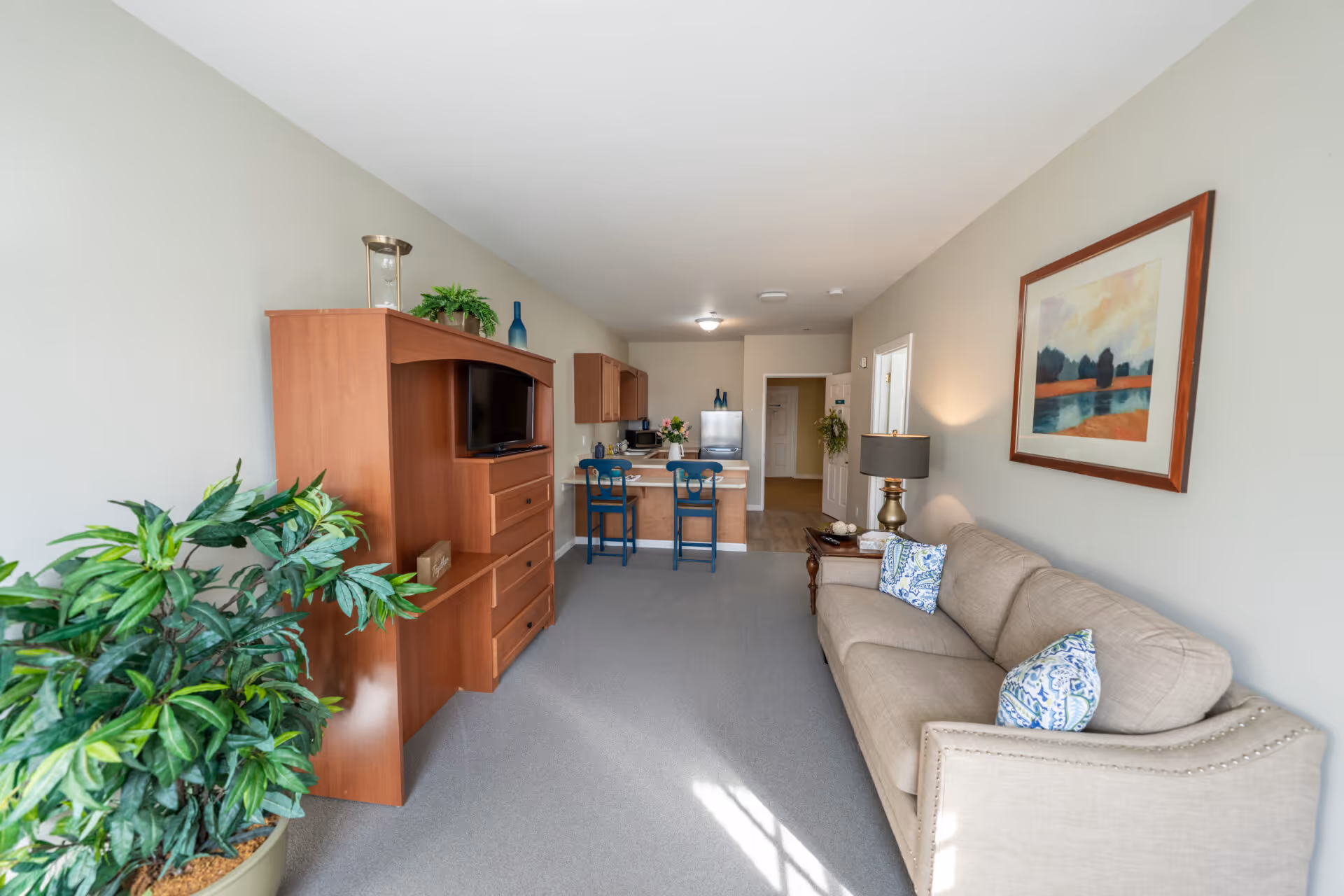 A bright living room area with a beige couch adorned with patterned pillows on the right, a wooden side table with a lamp, and a framed landscape painting on the wall above. On the left side, there is a large potted plant and a wooden entertainment center with a TV. In the background, a small kitchen area with a counter and two blue chairs is visible, along with a stainless steel refrigerator and cabinets.