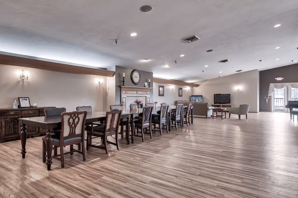 A spacious, well-lit common area with wooden flooring featuring a long dining table with chairs, a fireplace with a clock above it, a seating area with sofas and a TV, and large windows allowing natural light to enter.