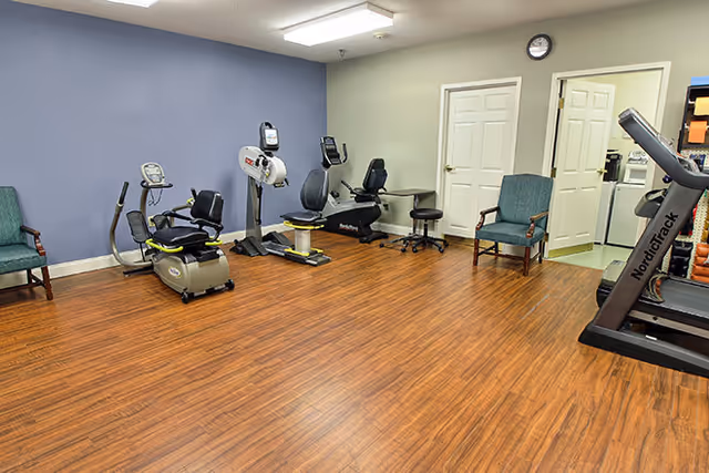 A fitness room with wooden flooring and light-colored walls, featuring exercise equipment including stationary bikes, a treadmill, and a stepper. There are two green upholstered chairs against the walls, a clock above two closed white doors, and a small black stool near the equipment.