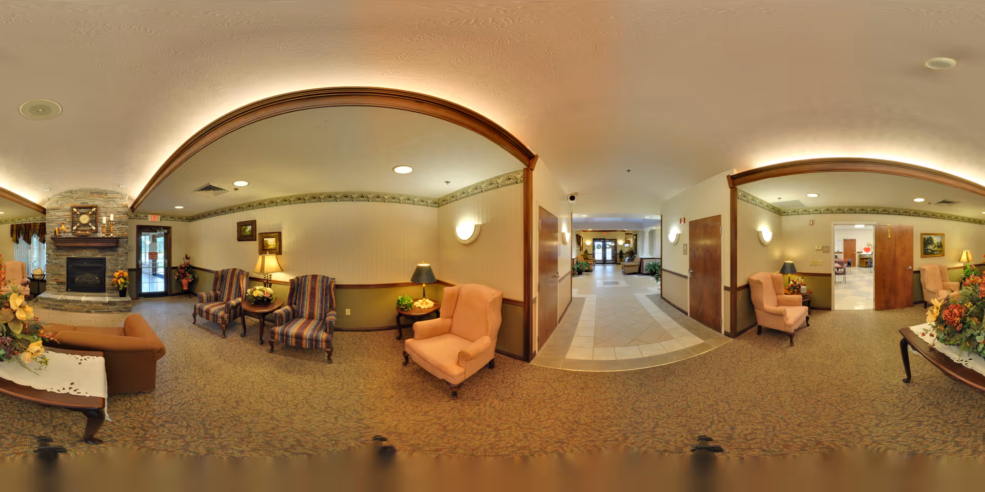 A spacious and warmly decorated senior living facility common area with comfortable upholstered chairs, side tables with lamps and floral arrangements, a stone fireplace, and a tiled hallway leading to an entrance door. The walls have a beige wallpaper with a decorative border near the ceiling, and the carpet is patterned in neutral tones.