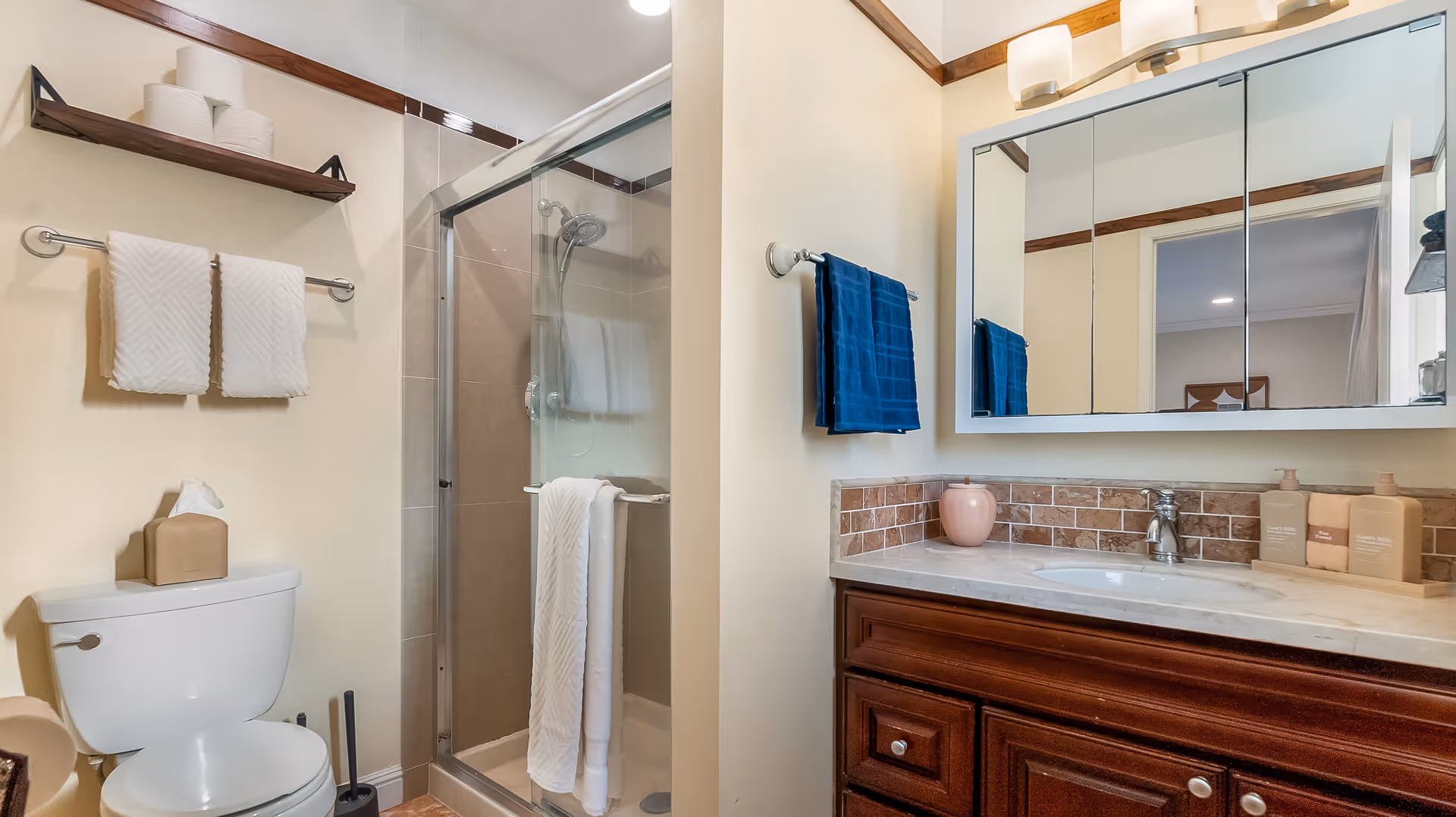 A clean and well-organized bathroom featuring a toilet with a tissue box on top, a glass-enclosed shower with a towel hanging on the door, a wooden shelf holding extra toilet paper rolls, and a vanity with a sink, a large mirror, and toiletries. The walls are light-colored with wooden trim accents.