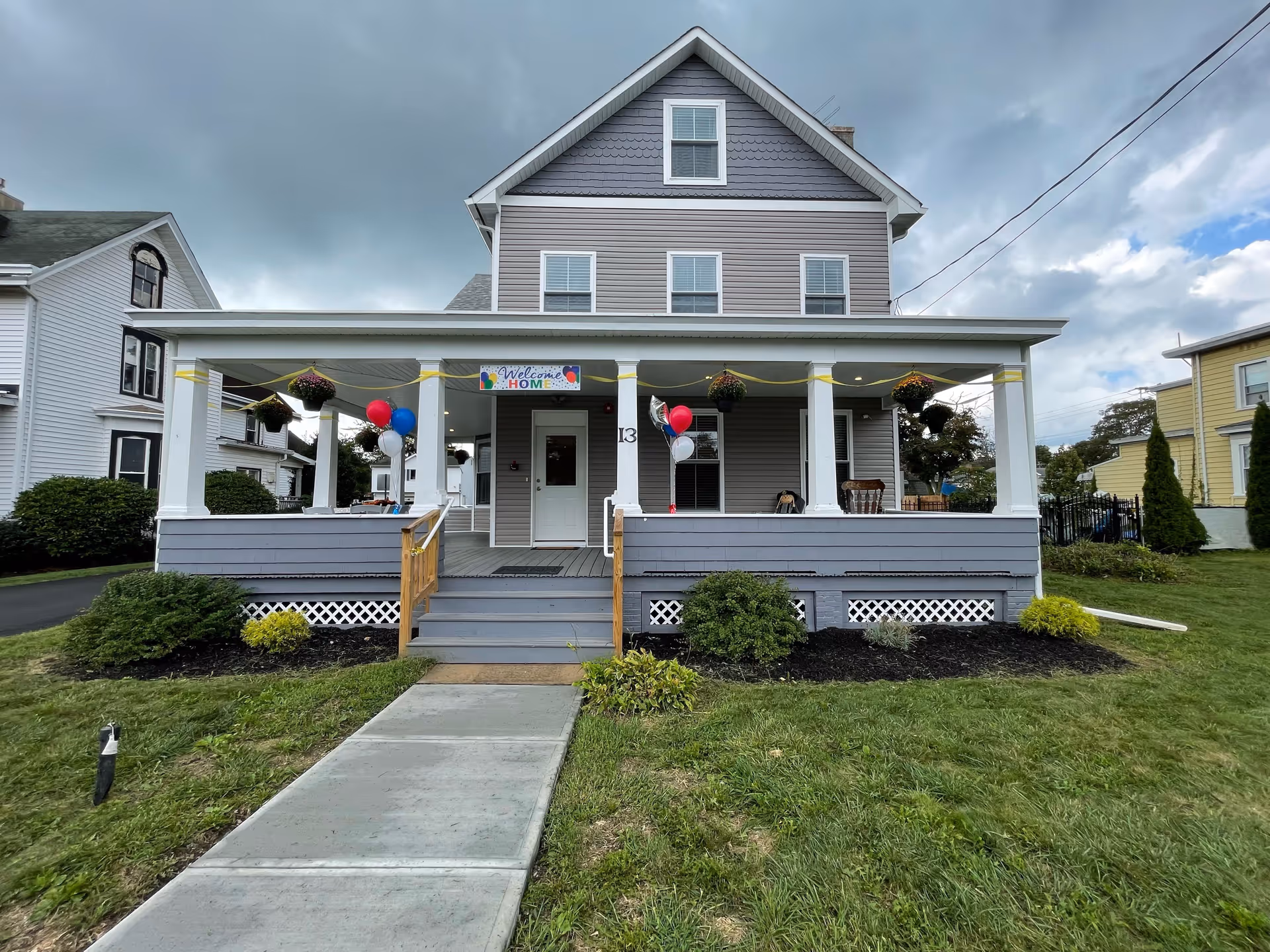 Front exterior view of a two-story gray house with a large porch decorated with hanging flower baskets, balloons, and a 'Welcome Home' banner. The house has white trim and a concrete walkway leading up to the porch steps. The sky is cloudy.