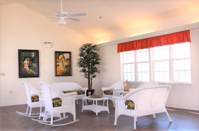 A bright and airy sitting area with white wicker furniture including two rocking chairs, two armchairs, and a loveseat, all with floral cushions. A small white wicker coffee table is in the center. The room has large windows with a red valance, two framed paintings on the wall, a tall potted plant in the corner, and a ceiling fan above.