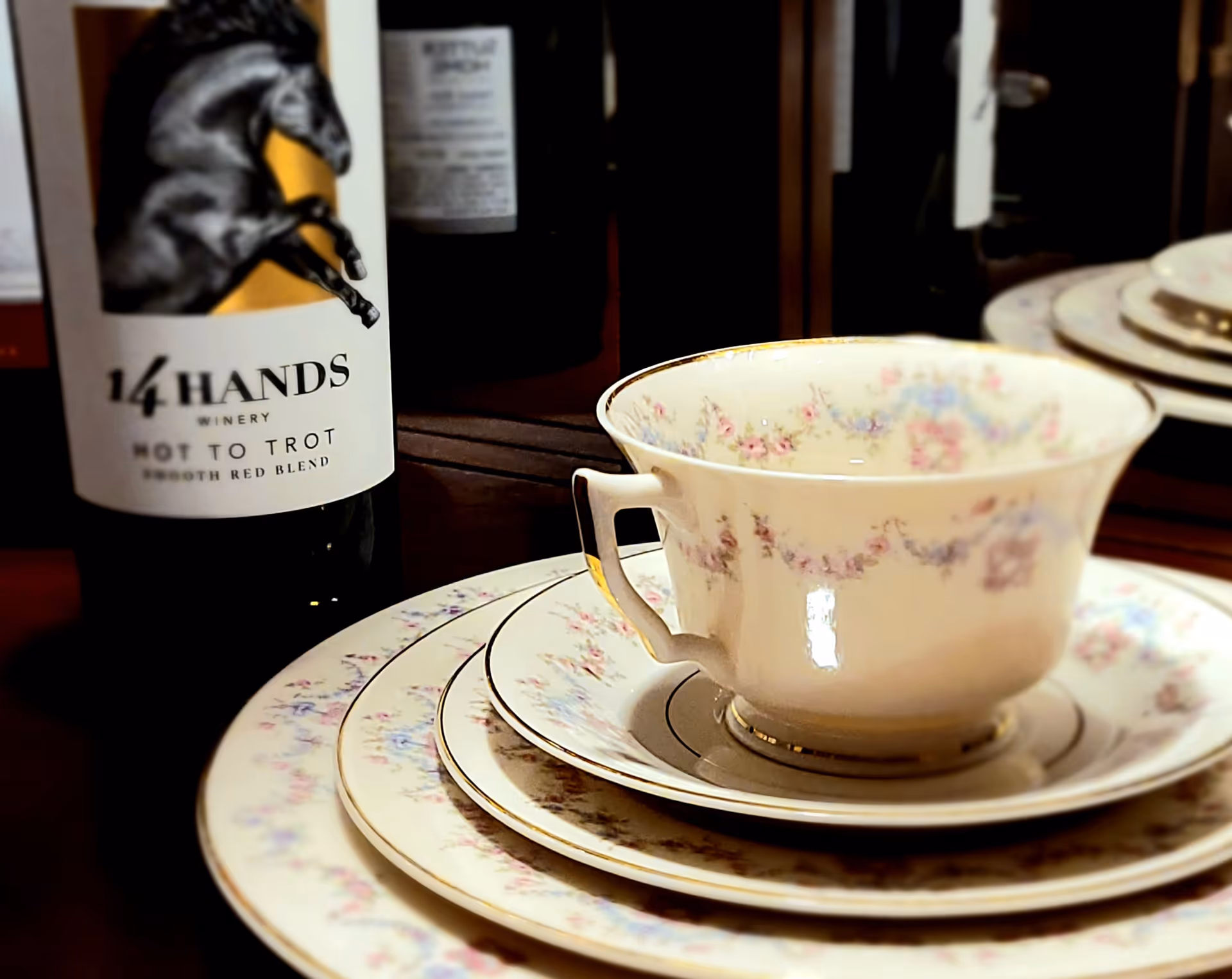 Close-up view of a set of floral-patterned china plates and a matching teacup with gold trim, placed on a table next to a bottle of 14 Hands Winery Hot to Trot Smooth Red Blend wine.