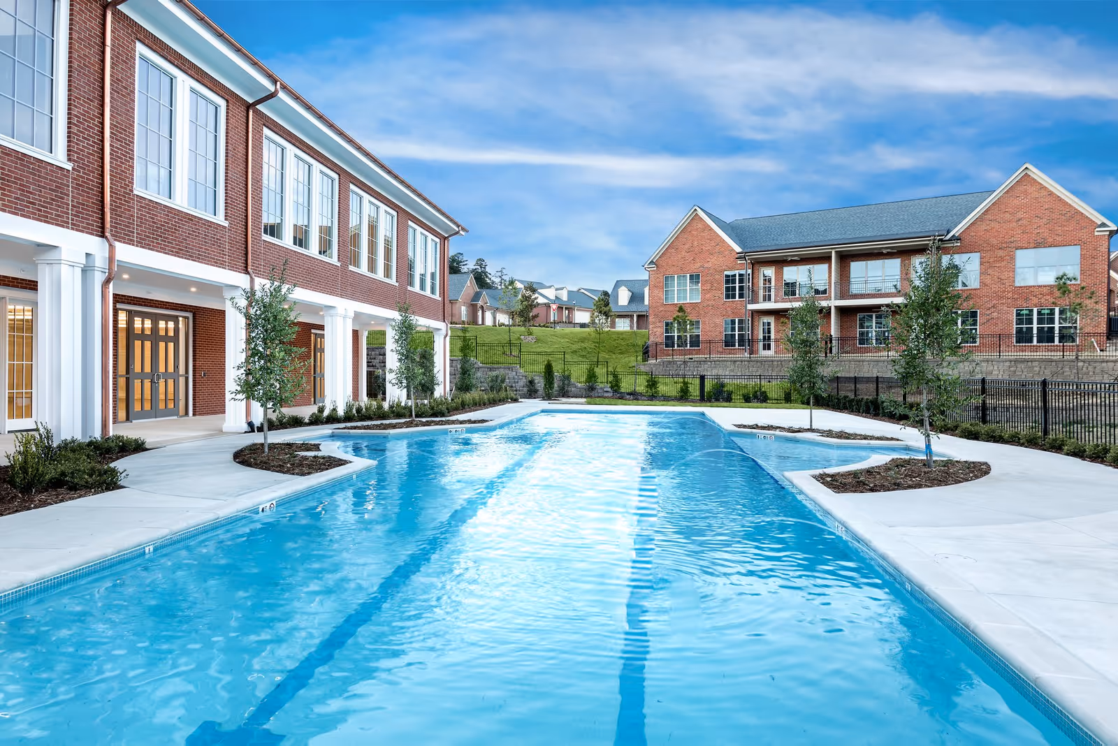 Outdoor swimming pool with clear blue water surrounded by a concrete deck and landscaped areas with small trees. Two large brick buildings with white columns and multiple windows are visible in the background under a blue sky with some clouds.