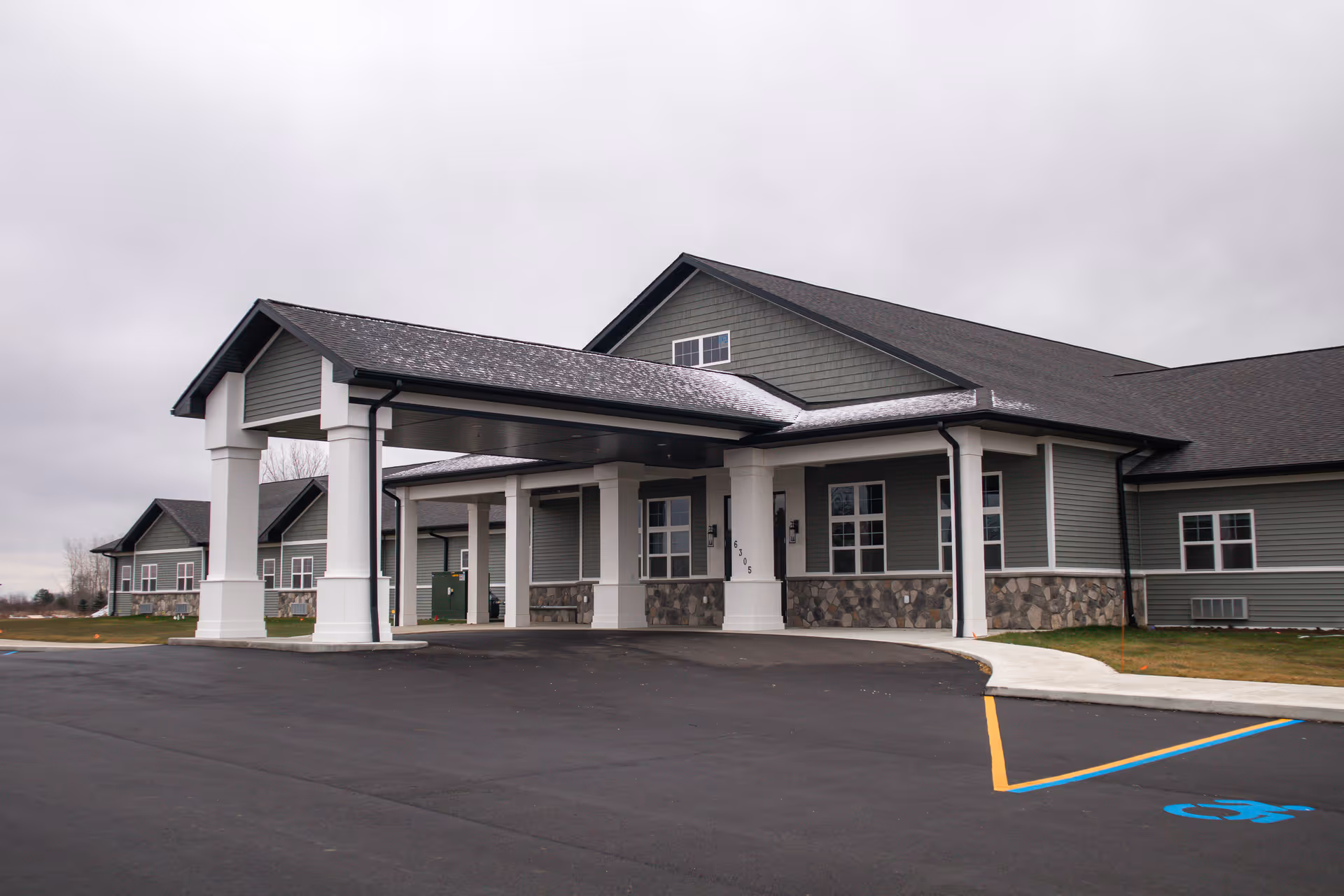 Exterior view of The Heartlands facility showing a large covered entrance with white pillars, gray siding, stone accents along the lower walls, and a paved driveway with a marked handicap parking space.