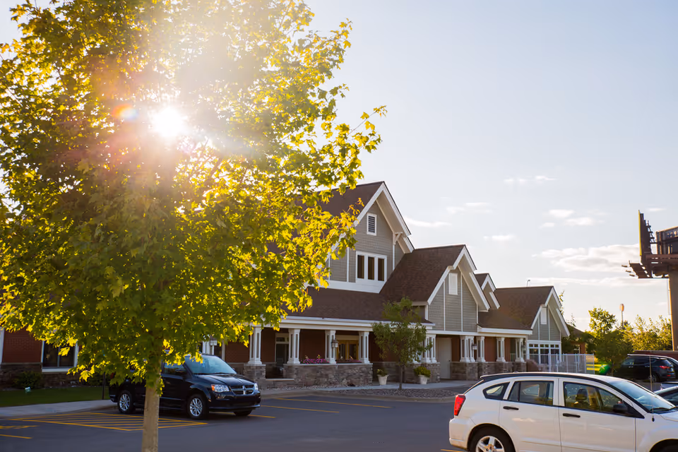 Exterior view of a senior living facility building with a tree in the foreground partially blocking the sun. Several cars are parked in the parking lot in front of the building under a clear sky.