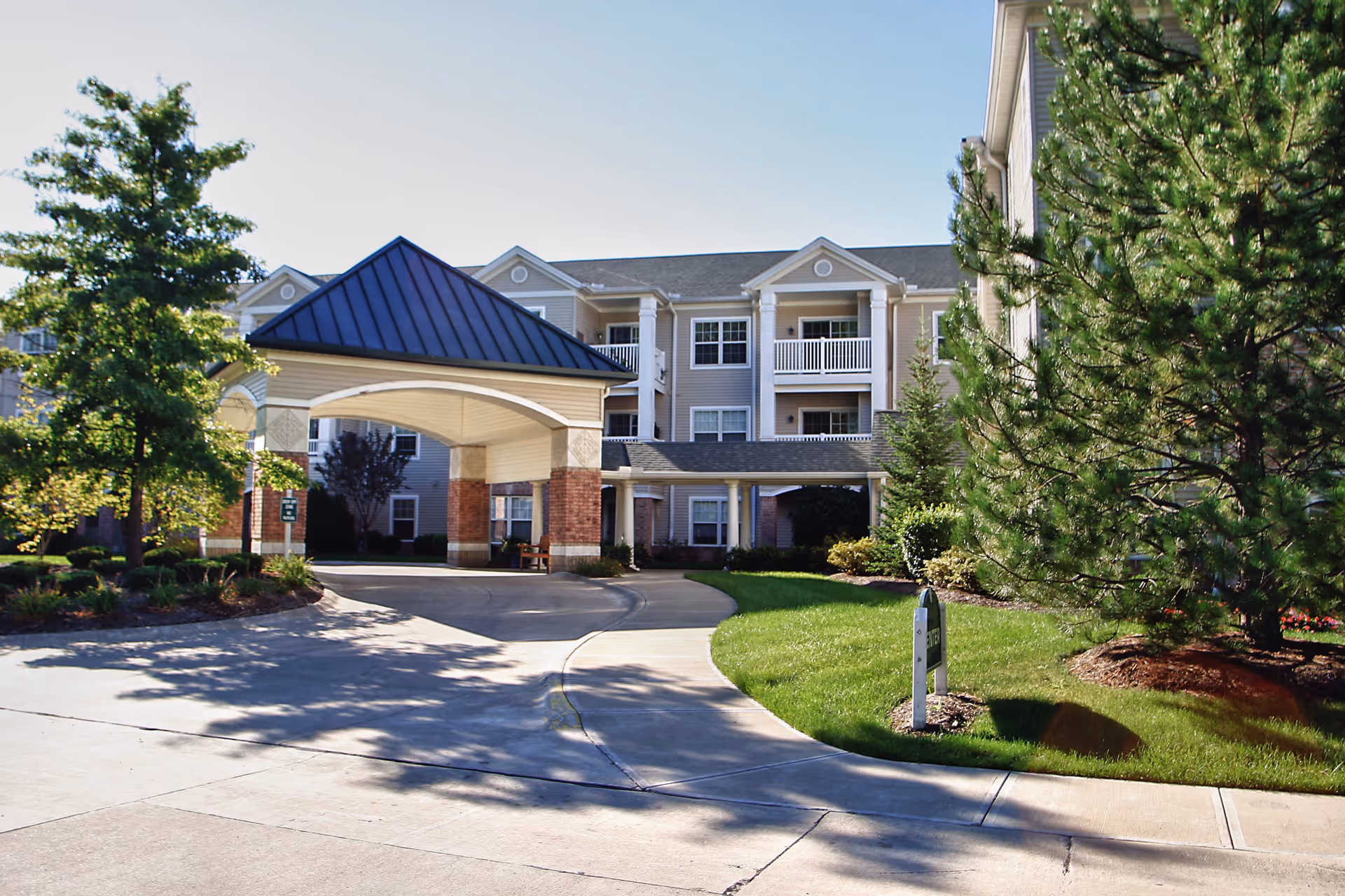 Exterior view of Commons Independent Living at Sprenger Health Care Towne Center Community Campus showing a covered driveway entrance with a peaked roof, surrounded by well-maintained landscaping including trees and shrubs, and a multi-story residential building in the background.