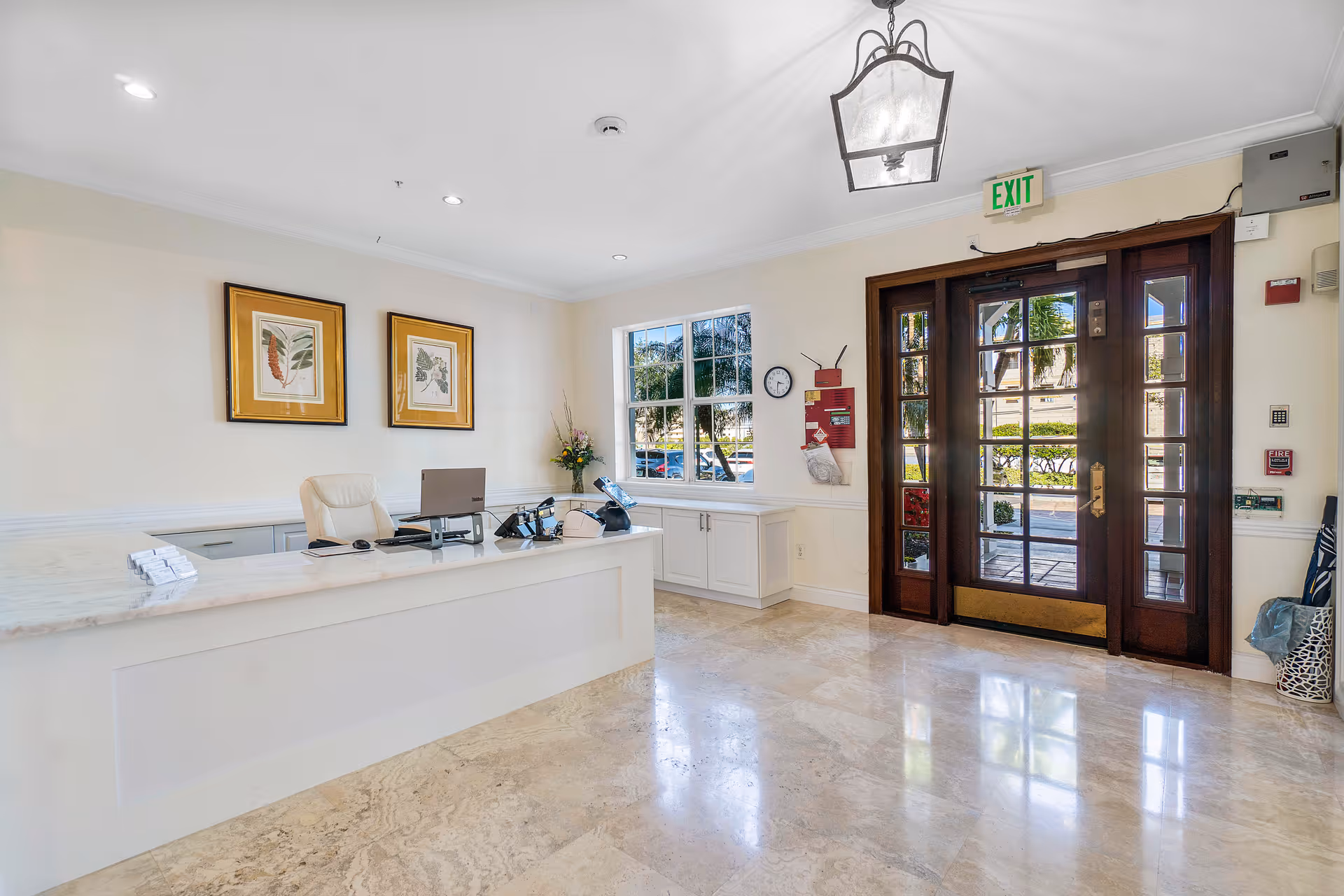Bright lobby reception area with a marble desk, office chair, framed artwork, window, and glass double entry doors.