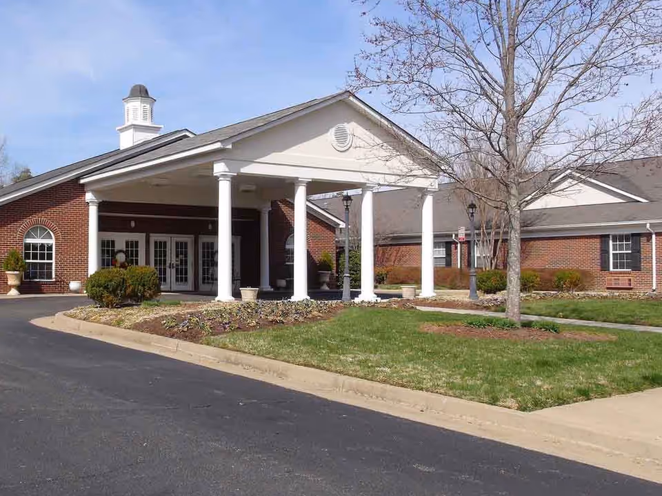 Exterior view of a senior living facility with a brick facade, white columns supporting a covered entrance, a driveway, landscaped garden beds, and a leafless tree on a sunny day.