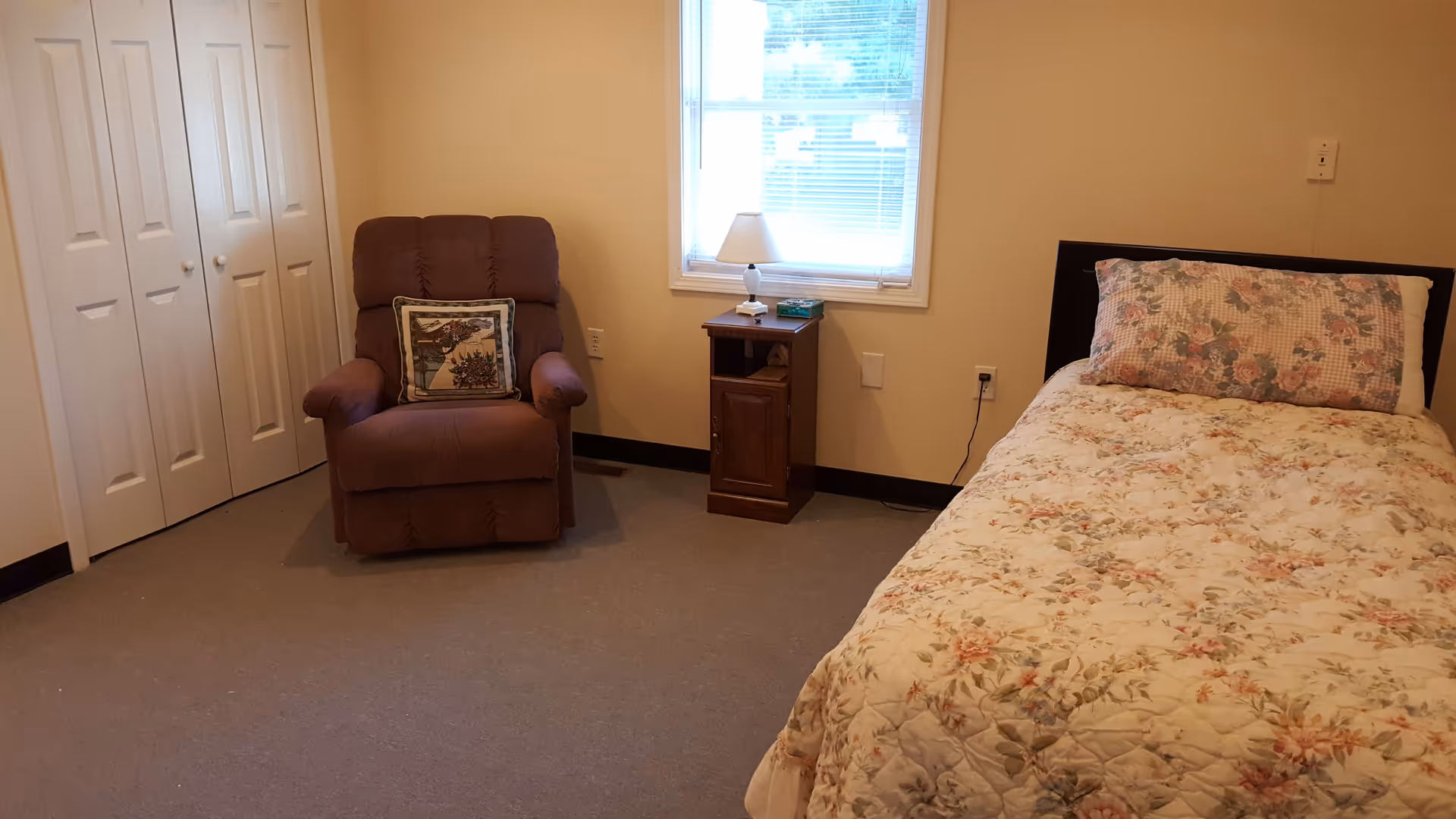 A simple bedroom with a single bed covered in a floral quilt and matching pillow. Next to the bed is a wooden nightstand with a small lamp and a box of tissues. A brown upholstered armchair with a decorative pillow is placed near a set of white double closet doors. A window with blinds lets in natural light.