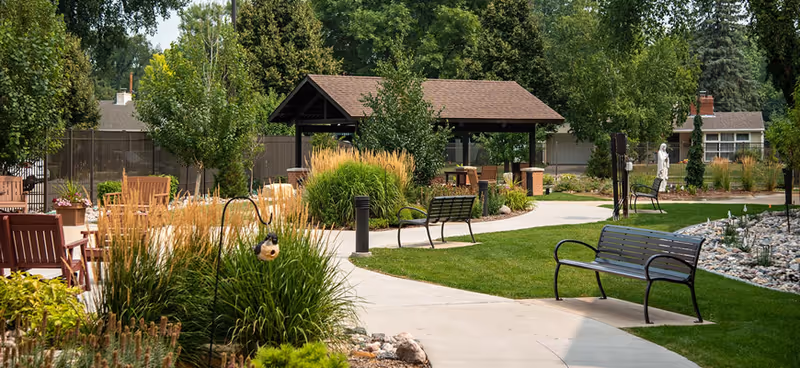 A peaceful outdoor garden area with a paved walkway, green grass, benches, ornamental grasses, and trees. There is a covered pavilion with seating in the background and a statue near a rock garden. Residential buildings and a wooden fence are visible in the background.
