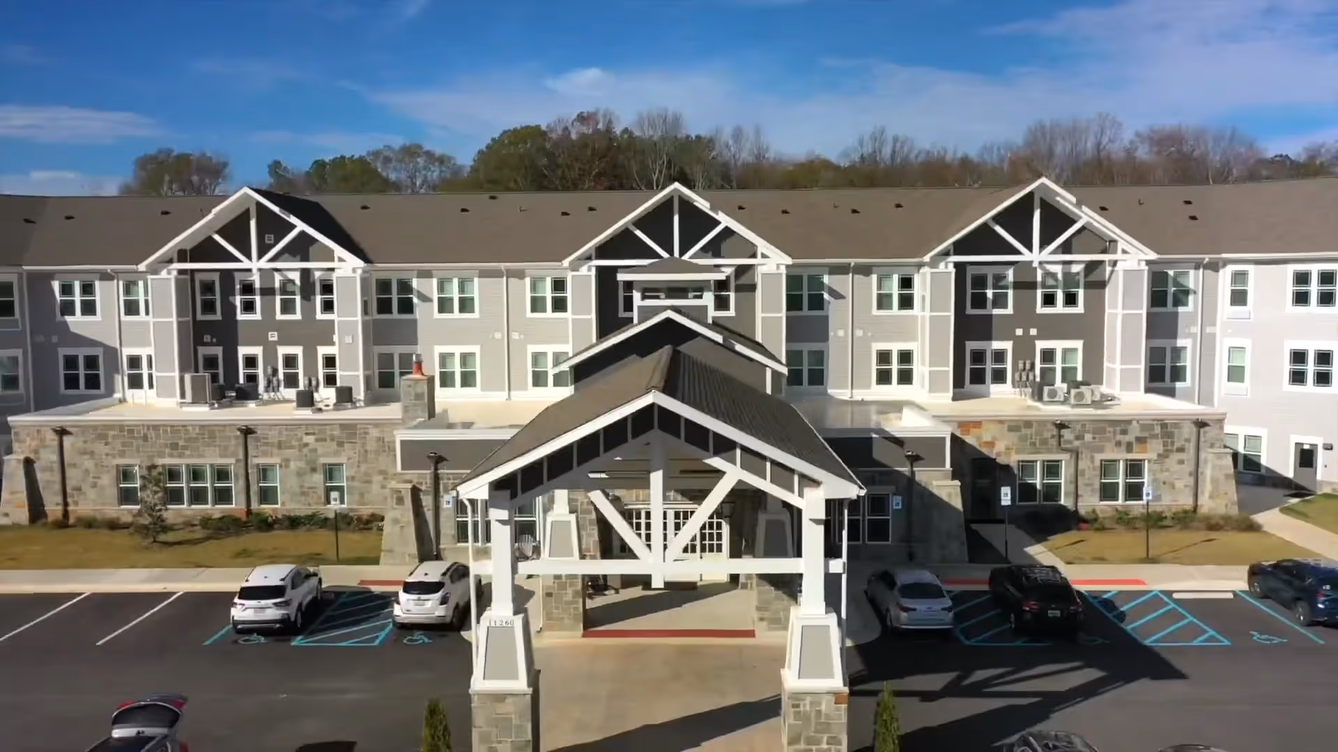 Front exterior view of a large, modern three-story senior living facility building with a covered entrance, multiple windows, and a parking lot with several cars parked in front. Trees and a blue sky with some clouds are visible in the background.
