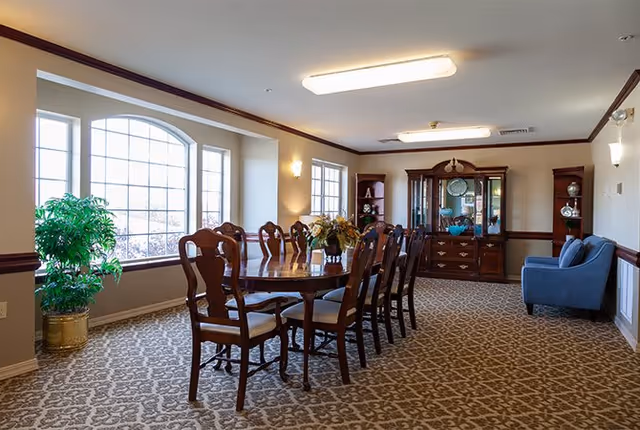 A furnished dining room with a long wooden table and chairs, a china cabinet, large window, and potted plant.