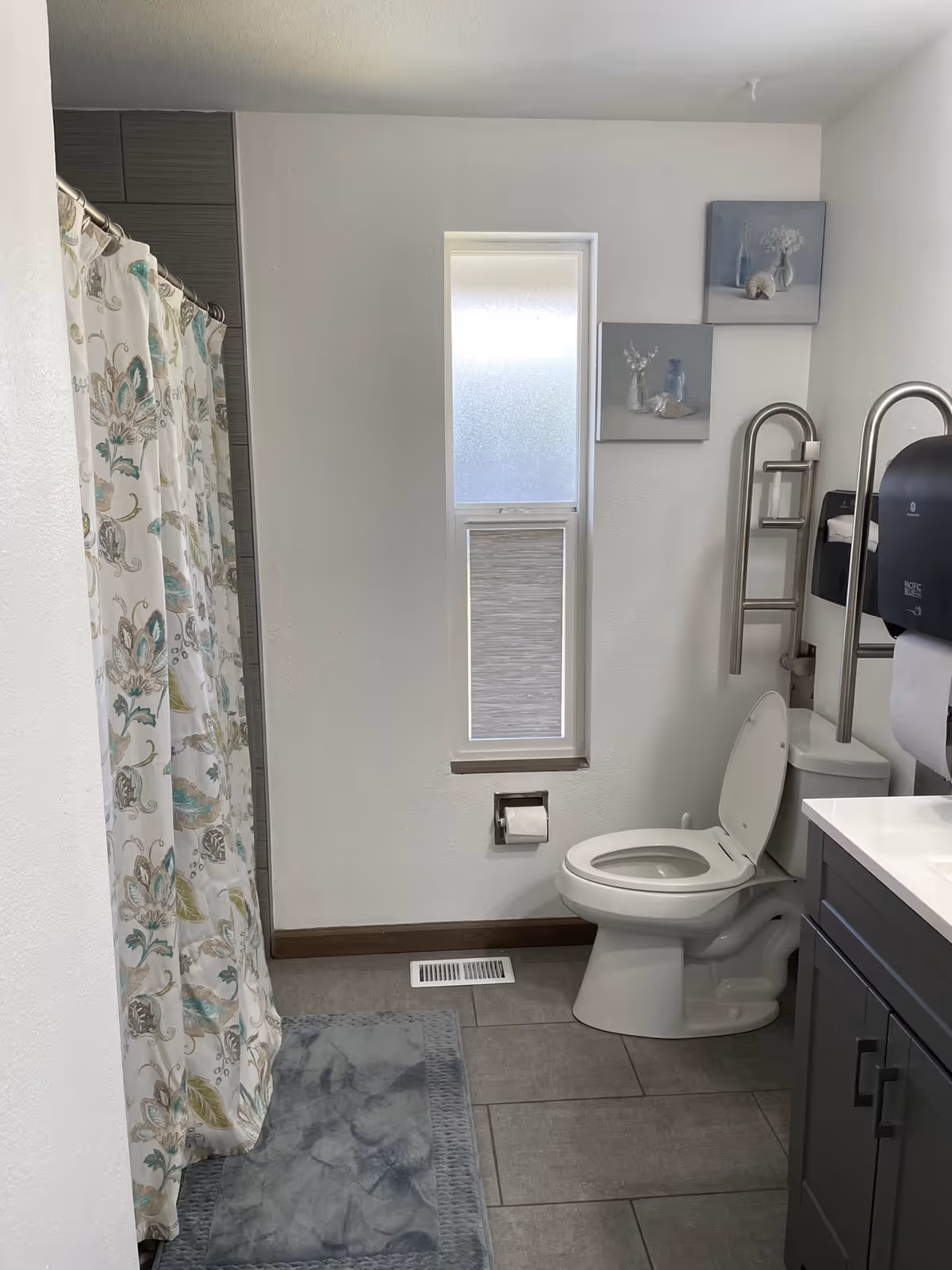 A clean bathroom with a toilet, a shower with a floral patterned curtain, a gray bath mat, a window with frosted glass and a blind, two small framed pictures on the wall, and a gray vanity with a white countertop. There are also metal grab bars near the toilet and a wall-mounted paper towel dispenser.