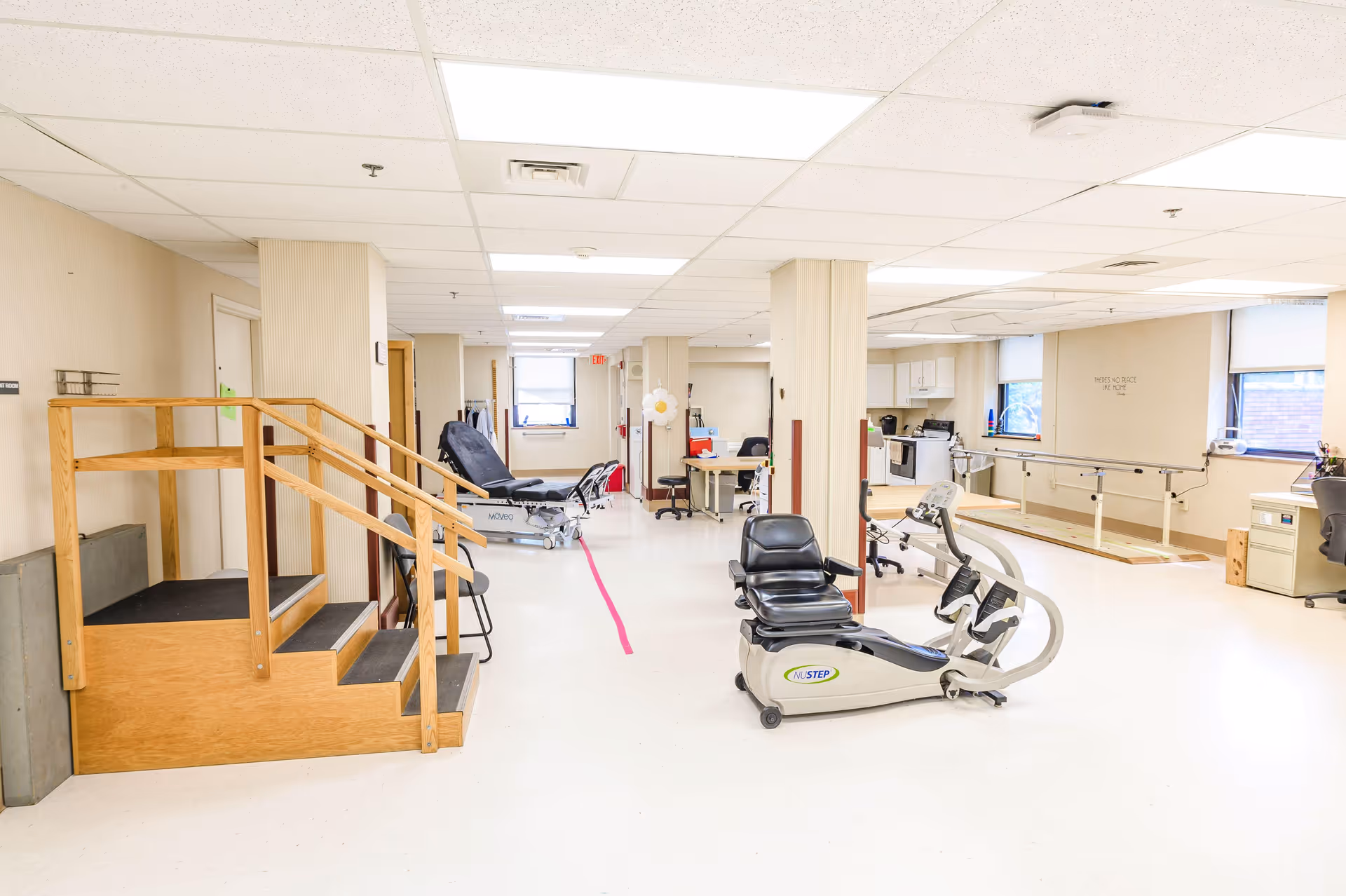 A bright rehabilitation room with exercise equipment including a NuStep recumbent cross trainer, parallel bars for walking practice, a wooden staircase for physical therapy, and several chairs and desks. The room has white walls, large windows, and a tiled ceiling with fluorescent lighting.