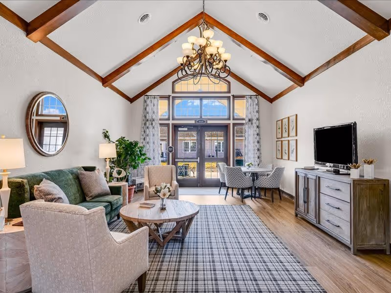 A bright and spacious living room with a high vaulted ceiling featuring wooden beams and a chandelier. The room includes a green sofa, two beige armchairs, a round wooden coffee table with a flower arrangement, a plaid area rug, a wooden TV console with a flat-screen TV, and a small dining table with four chairs near large windows and glass doors letting in natural light. The walls are decorated with a round mirror and framed artwork.