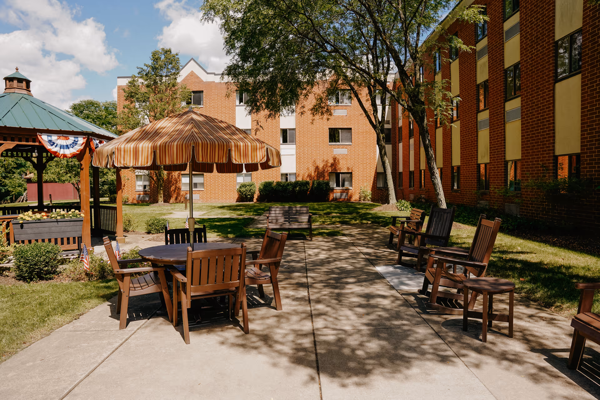 Outdoor seating area at a senior living facility with wooden chairs and tables, one table shaded by a striped umbrella. A gazebo decorated with patriotic bunting is visible on the left, surrounded by green grass, trees, and brick buildings in the background under a partly cloudy sky.